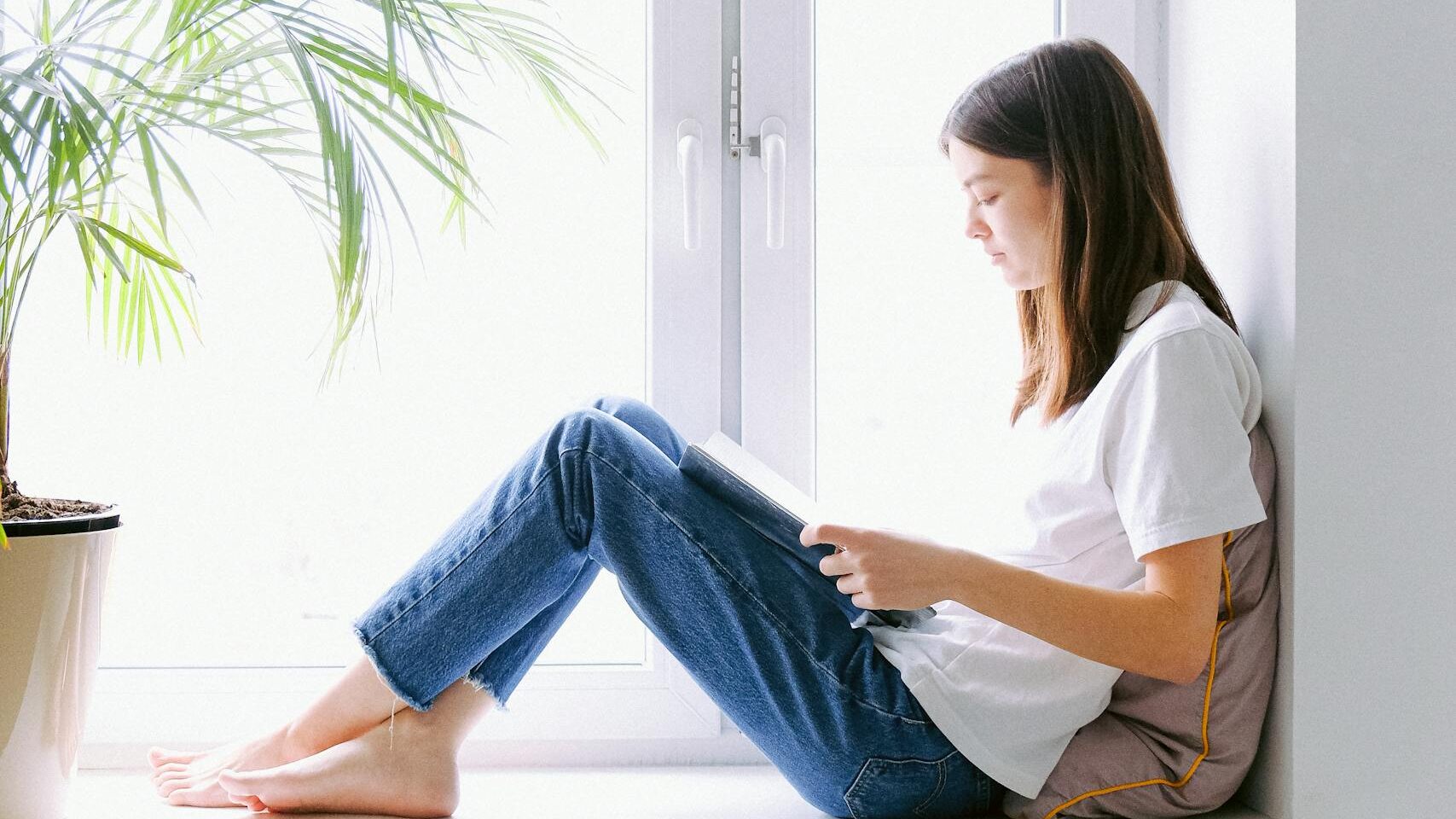 Teen girl enjoying a book while sitting by a bright window indoors.